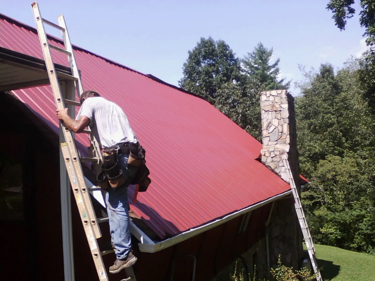 Roofer on a ladder beside a red metal roof and stone chimney — Soffit & Fascia Repair by Roofing Brothers in Roslindale